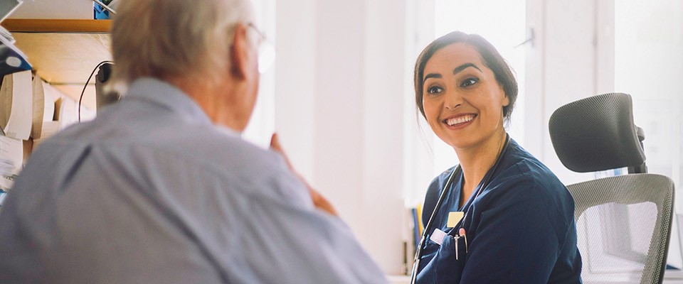 Eine freundliche Ärztin oder Pflegekraft in blauer medizinischer Kleidung mit Stethoskop um den Hals sitzt an einem Schreibtisch und spricht mit einem älteren Patienten. Sie lächelt ihn an, während er ihr aufmerksam zuhört. Der Hintergrund zeigt eine helle Arztpraxis mit einem Fenster. Auf der rechten Seite des Bildes befindet sich eine grüne Grafik mit einem Symbol von einer Lunge.