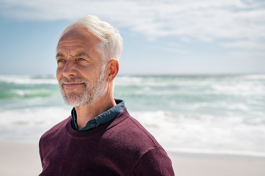 Das Bild zeigt einen älteren Mann, der am Strand mit dem Rücken zum Meer steht und in die Ferne blickt.