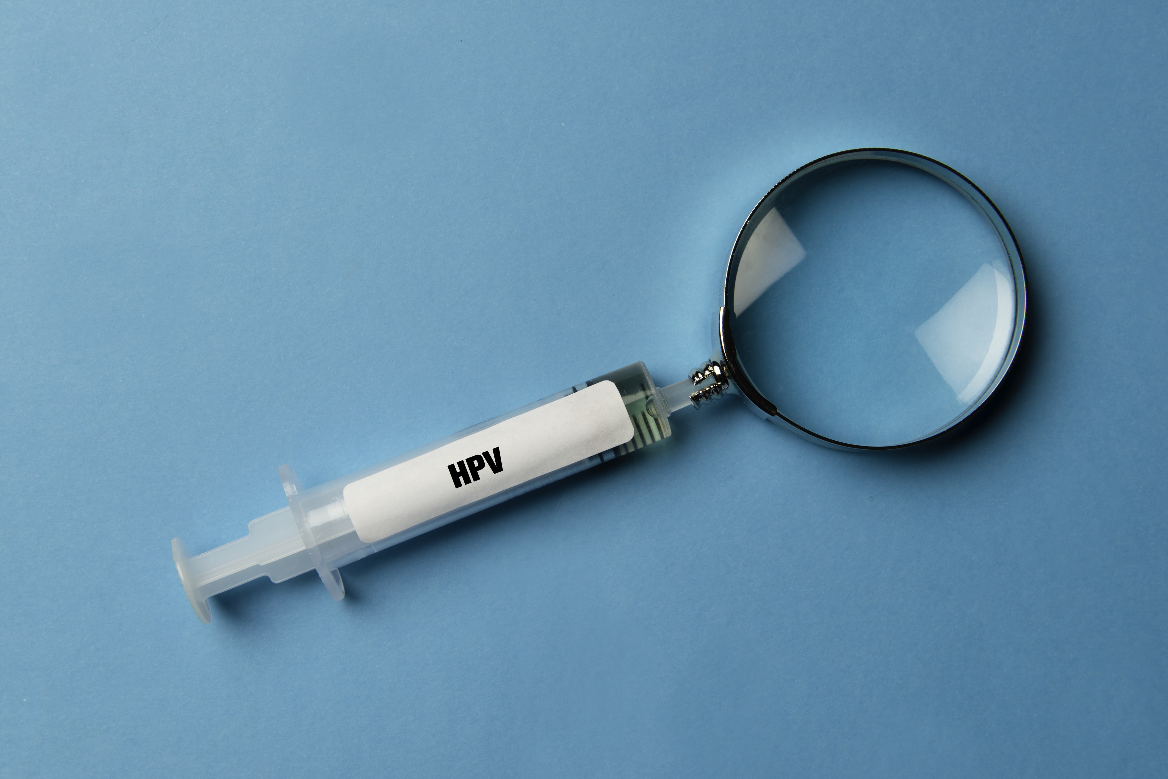 Impfspritze mit dem Aufdruck "HPV". Statt der Impfnadel hat die Spritze eine Lupe am oberen Ende.