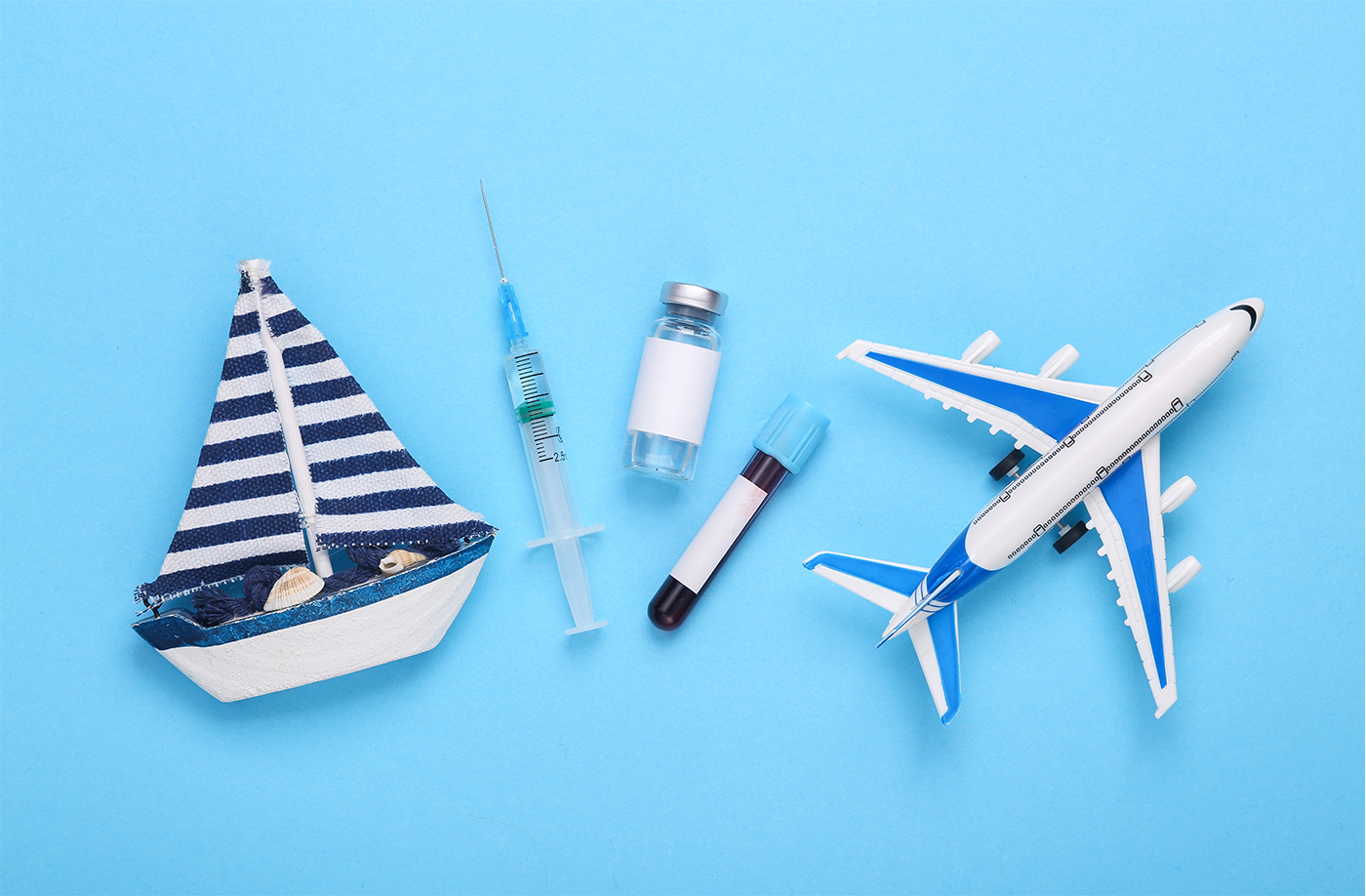 Spielzeug Segelboot, Spritze, Fläschchen und ein Spielzeug Flugzeug liegen nebeneinander auf blauem Untergrund.