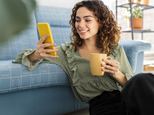 Eine junge Frau mit lockigem Haar sitzt lächelnd auf dem Boden vor einem Sofa, hält eine gelbe Tasse in der einen und ein gelbes Smartphone in der anderen Hand. Sie trägt eine grüne Bluse mit Rüschen und scheint entspannt eine Nachricht zu lesen oder ein Video anzusehen. Die Szene wirkt freundlich und gemütlich, aufgenommen in einem modernen Wohnzimmer.