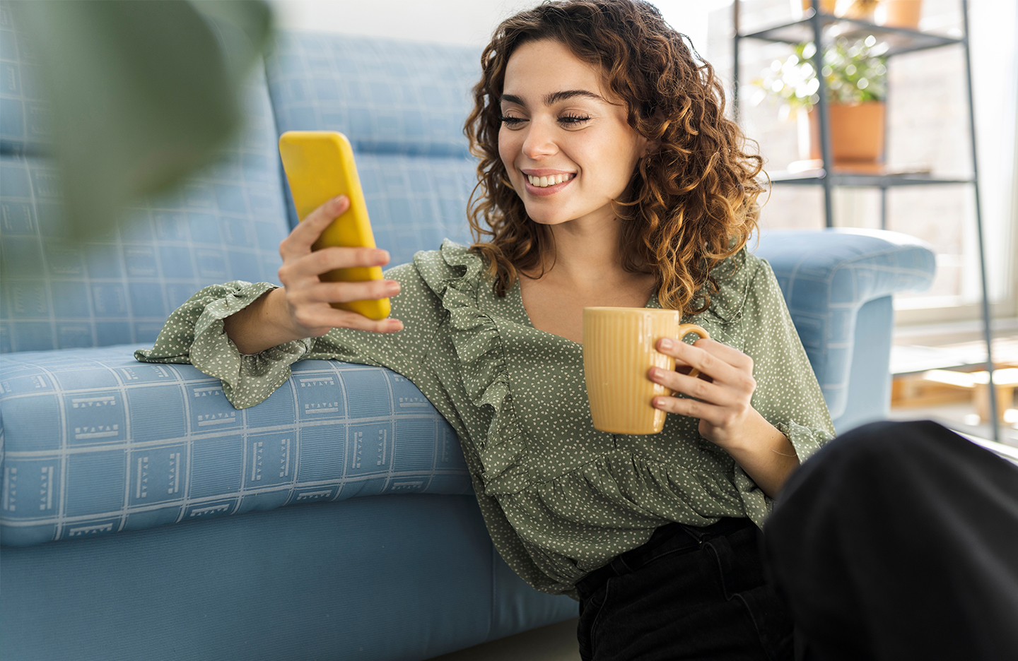 Eine junge Frau mit lockigem Haar sitzt lächelnd auf dem Boden vor einem Sofa, hält eine gelbe Tasse in der einen und ein gelbes Smartphone in der anderen Hand. Sie trägt eine grüne Bluse mit Rüschen und scheint entspannt eine Nachricht zu lesen oder ein Video anzusehen. Die Szene wirkt freundlich und gemütlich, aufgenommen in einem modernen Wohnzimmer.