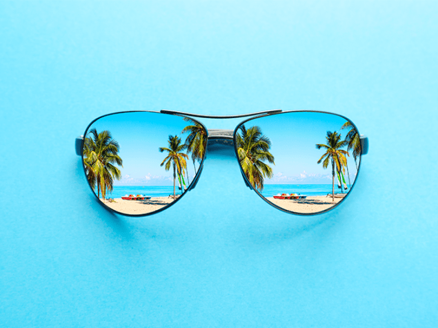 Die Abbildung zeigt eine große Sonnenbrille vor einem hellblauen Hintergrund. In den Gläsern spiegeln sich ein Strand mit Palmen und Sand.
