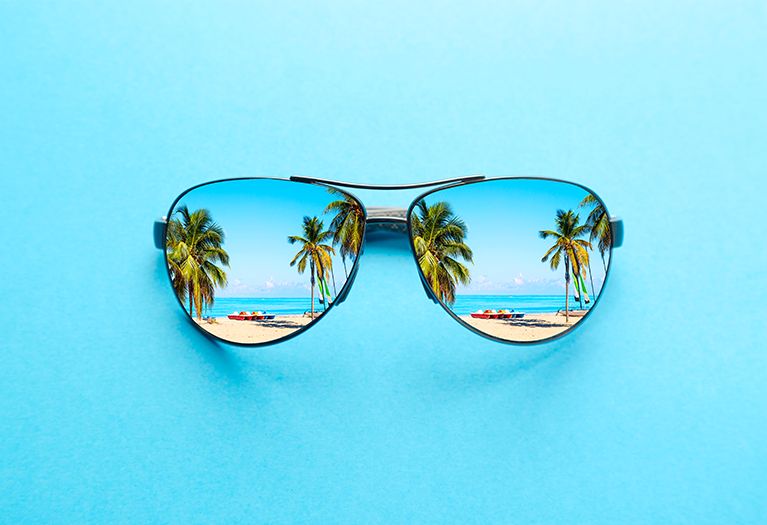 Die Abbildung zeigt eine große Sonnenbrille vor einem hellblauen Hintergrund. In den Gläsern spiegeln sich ein Strand mit Palmen und Sand.
