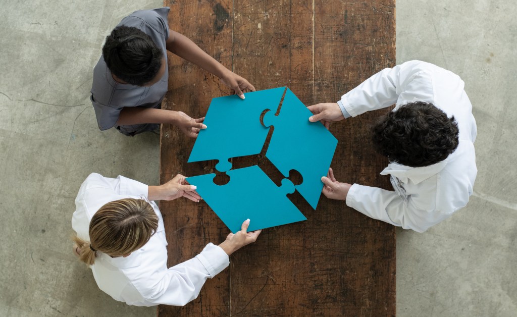 Drei Personen, darunter zwei in weißen Kitteln, stehen um einen Holztisch und fügen gemeinsam vier große, blaue Puzzleteile zusammen. Die Szene ist aus der Vogelperspektive aufgenommen und symbolisiert Zusammenarbeit, Teamwork und gemeinsames Problemlösen – möglicherweise im medizinischen oder wissenschaftlichen Kontext.
