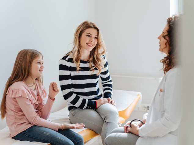 Ein junges Mädchen und ihre Mutter sitzen lächelnd in einem hellen Behandlungszimmer auf einer Untersuchungsliege und führen ein Gespräch mit einer Ärztin, die ihnen gegenüber sitzt. Die Ärztin trägt einen weißen Kittel und hört aufmerksam zu. Die Szene vermittelt eine freundliche, offene und vertrauensvolle Atmosphäre in einem Arzt-Patienten-Gespräch.