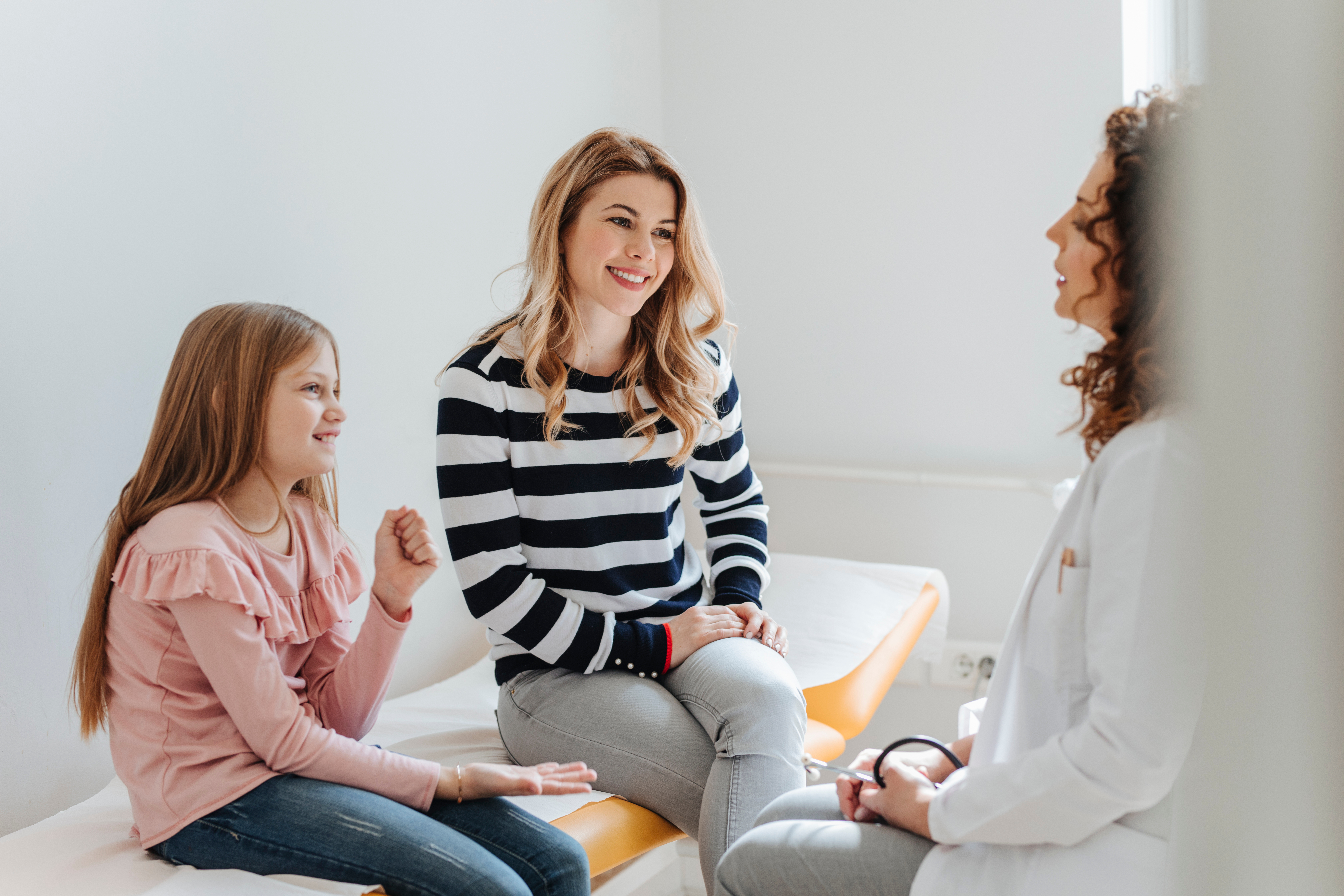 Ein junges Mädchen und ihre Mutter sitzen lächelnd in einem hellen Behandlungszimmer auf einer Untersuchungsliege und führen ein Gespräch mit einer Ärztin, die ihnen gegenüber sitzt. Die Ärztin trägt einen weißen Kittel und hört aufmerksam zu. Die Szene vermittelt eine freundliche, offene und vertrauensvolle Atmosphäre in einem Arzt-Patienten-Gespräch.