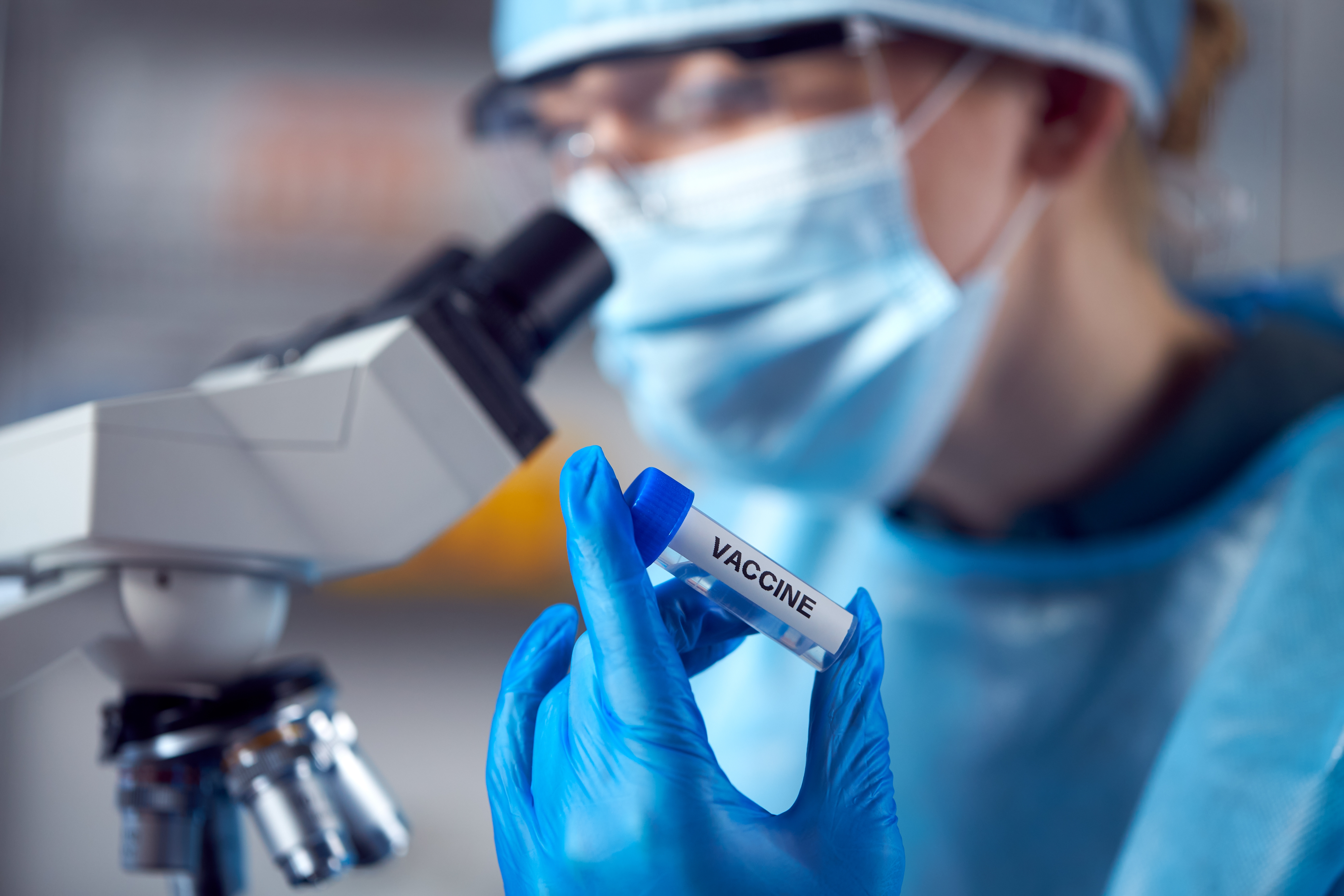 Eine Person im Labor mit blauen medizinischen Handschuhen hält ein Impfstoffviol in den Vordergrund. Dabei schaut sie in ein Mikroskop
