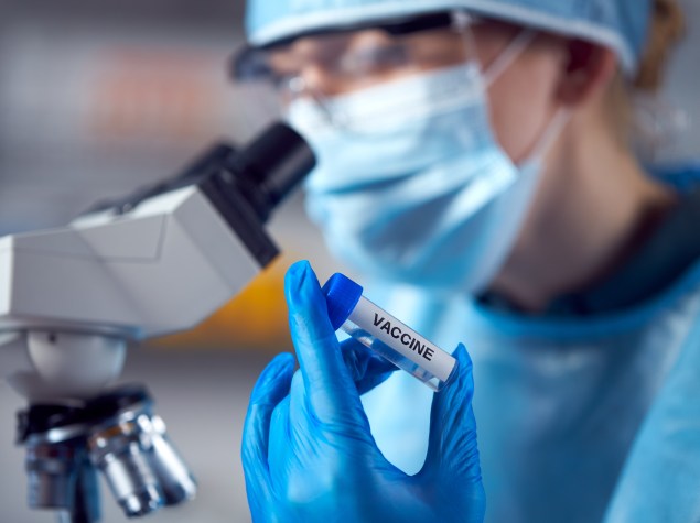 Eine Person im Labor mit blauen medizinischen Handschuhen hält ein Impfstoffviol in den Vordergrund. Dabei schaut sie in ein Mikroskop