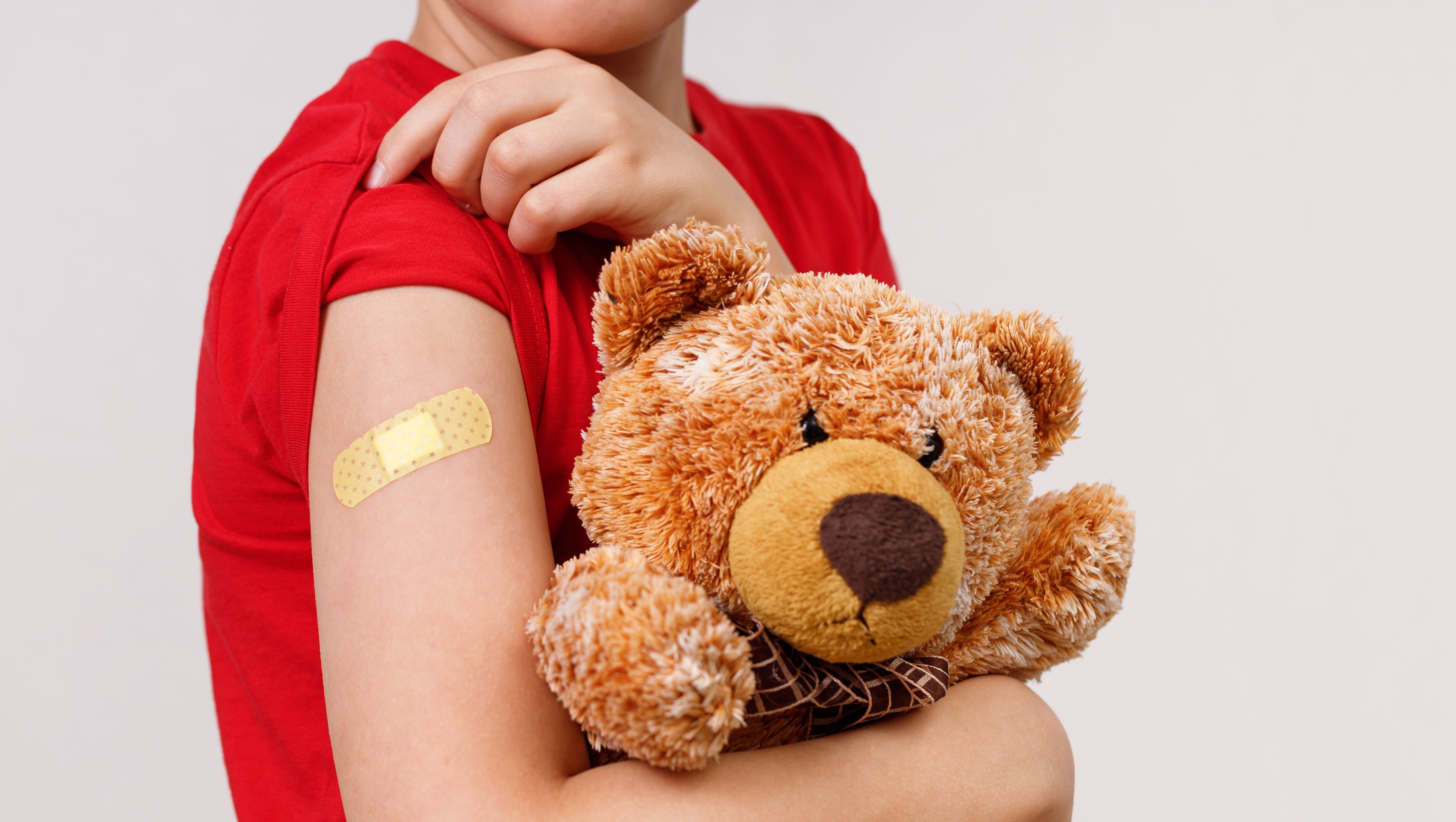 Kind zeigt Pflaster auf dem Arm und hält einen Teddy. 