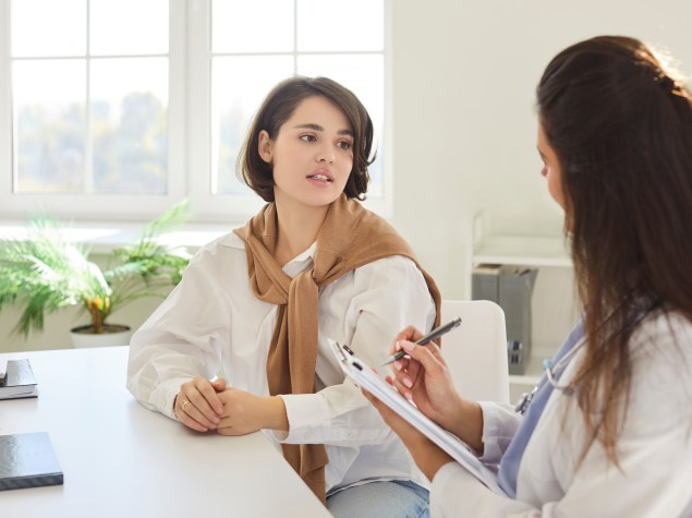 Eine Patientin sitzt an einem Tisch und spricht mit einer Ärztin, die Notizen auf einem Klemmbrett macht. Beide sind einander zugewandt und wirken in ein konzentriertes Gespräch vertieft. Die Umgebung ist hell und freundlich mit einem Fenster und Pflanzen im Hintergrund