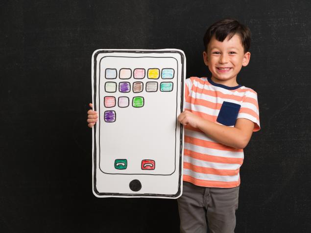 Ein lächelnder Junge steht vor dunklem Hintergrund und hält eine große Bastelvorlage in Form eines Smartphones mit bunten gezeichneten App-Symbolen.